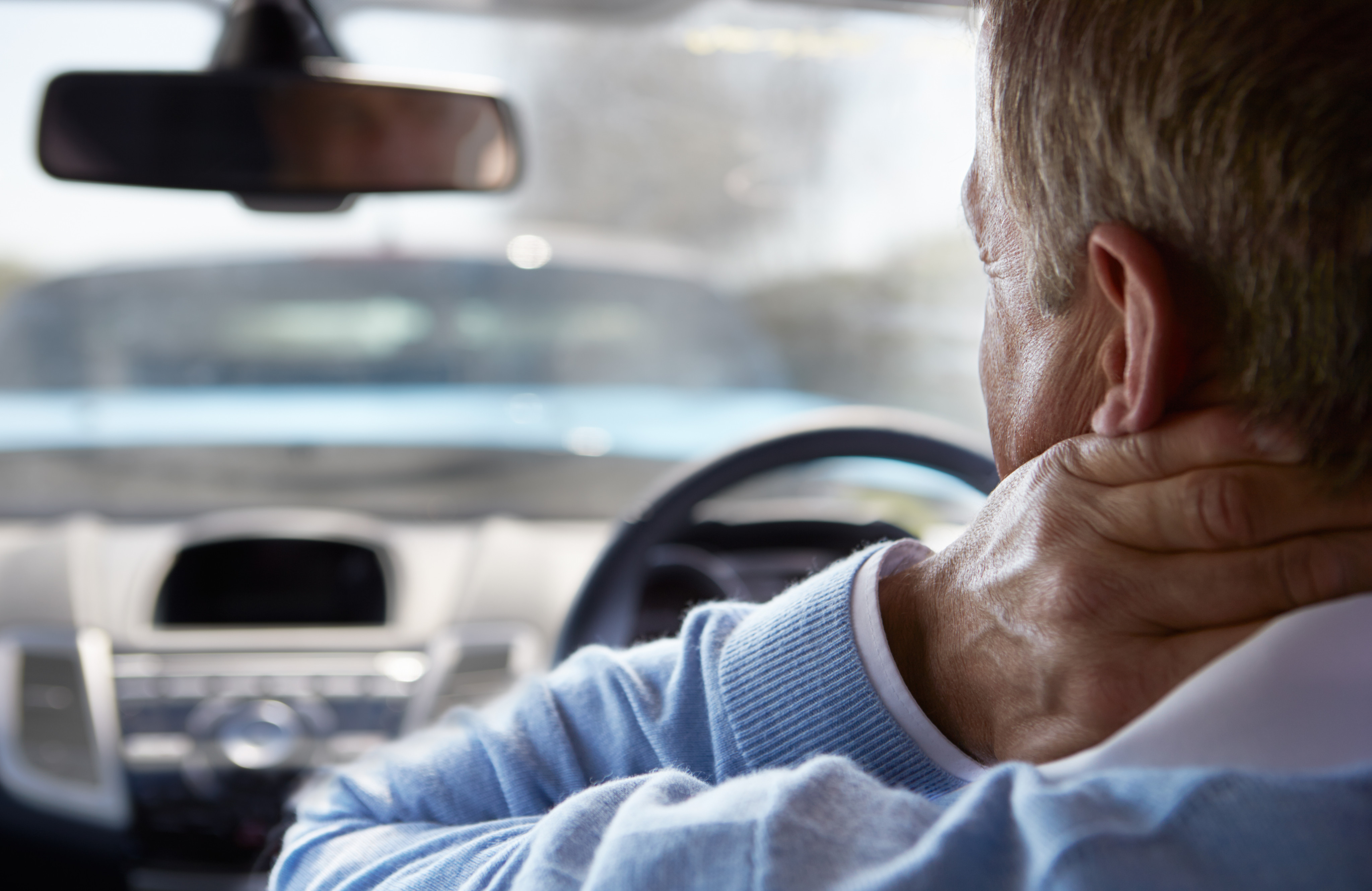 man holding the back of his neck in pain behind the steering wheel of a car